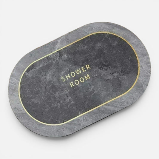 Clairly oval absorbent diatomite bath mat with a grey marble print and gold Shower Room text on a tiled floor.