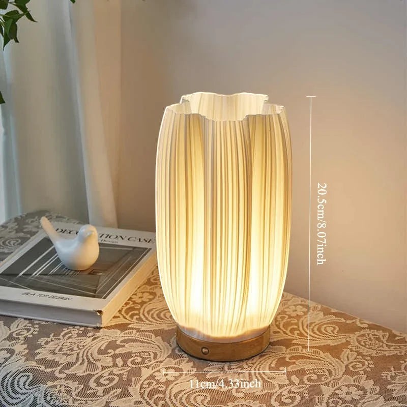 Clairly sculptural desk lamp with a pleated ivory shade and solid wood base glowing on a patterned table cloth