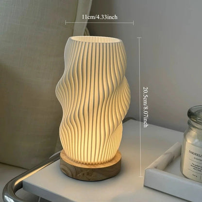 Clairly sculptural desk lamp with a pleated ivory shade and solid wood base, emitting a warm glow on a nightstand.