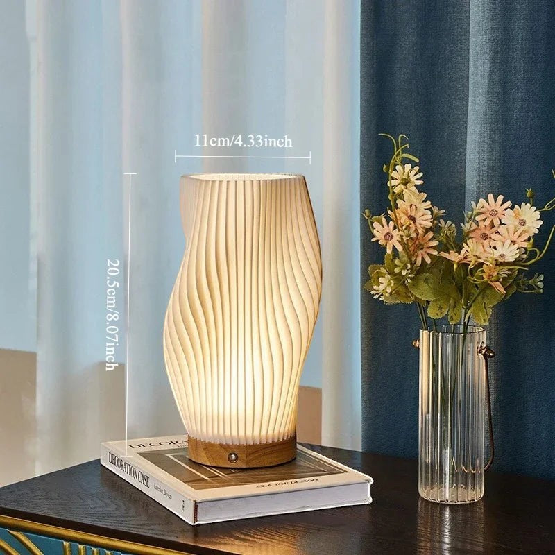 Clairly sculptural desk lamp with a pleated white shade and solid wood base glowing on a bedside table.