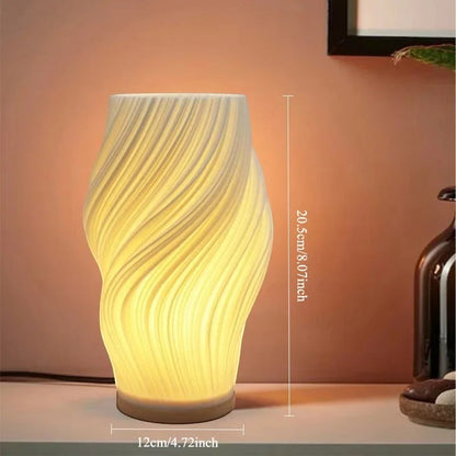 Clairly sculptural desk lamp with a white pleated shade and solid wood base emitting a warm glow on a table