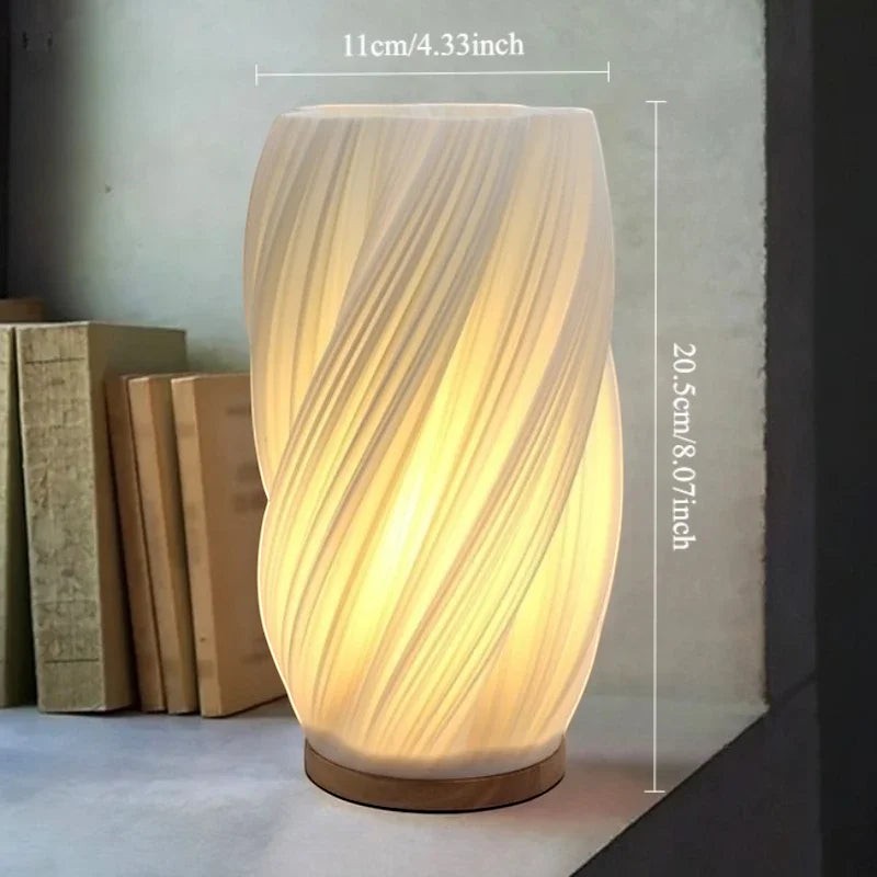Clairly sculptural desk lamp with a twisted pleated shade and solid wood base emitting a warm glow on a shelf.