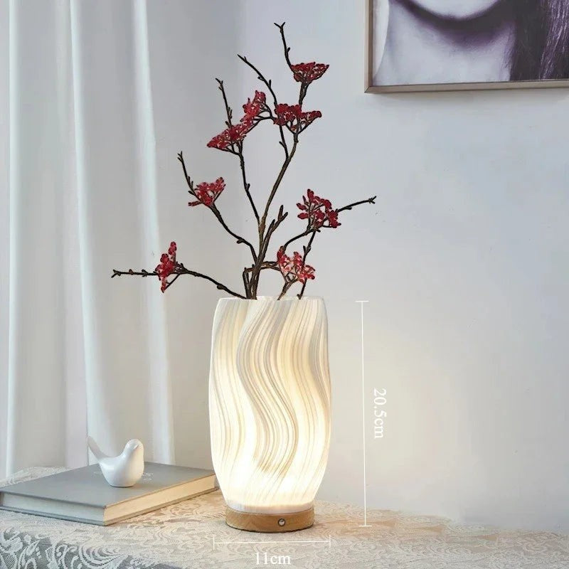 Clairly sculptural desk lamp with a white pleated shade and solid wood base, glowing with warm light on a table.